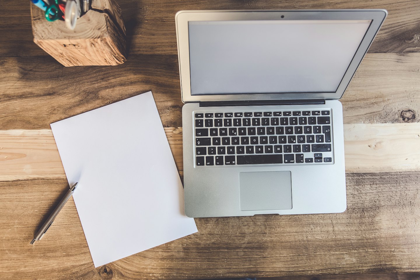 Silver laptop on a wooden desk with paper and pens.